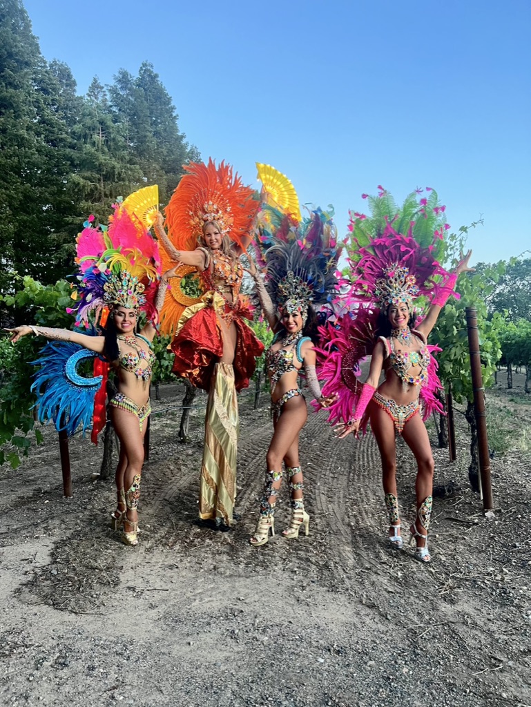 Stilt performer in an orange feathered costume with three colorful carnival performers