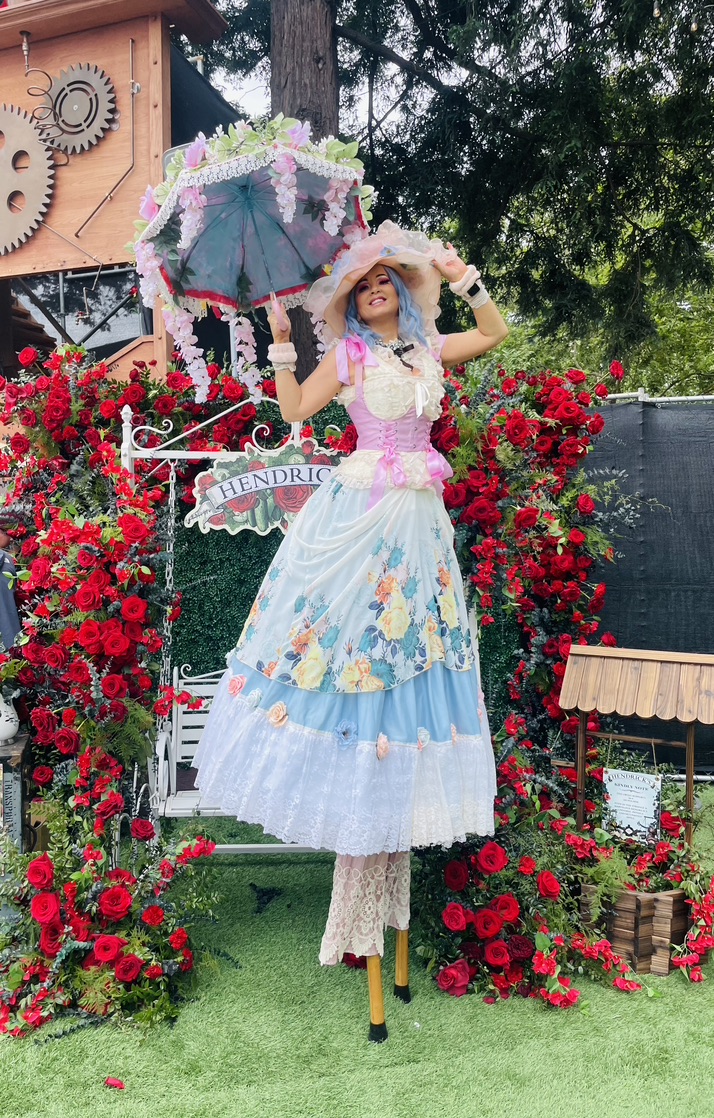 Miss Molly on stilts in a pastel floral dress holding a flower-trimmed parasol