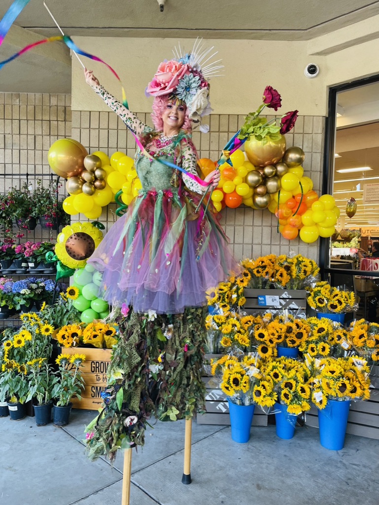 Miss Molly on stilts waving ribbons beside a flower display