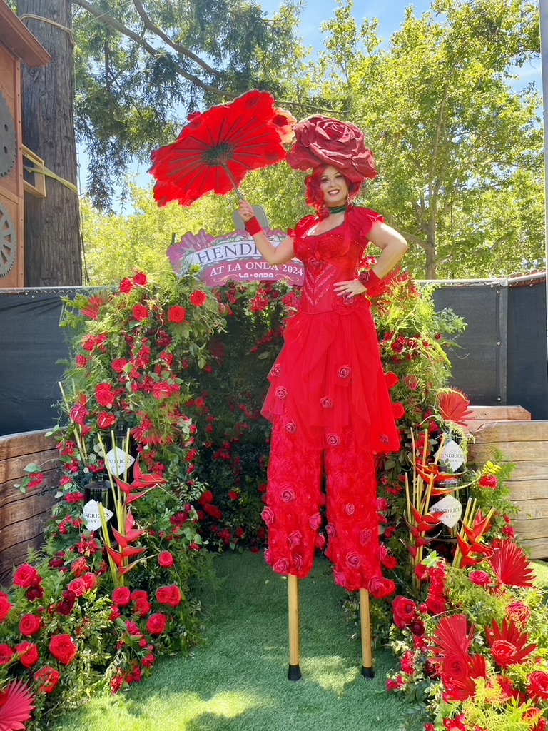 Miss Molly on stilts wearing a red rose costume and holding a red parasol