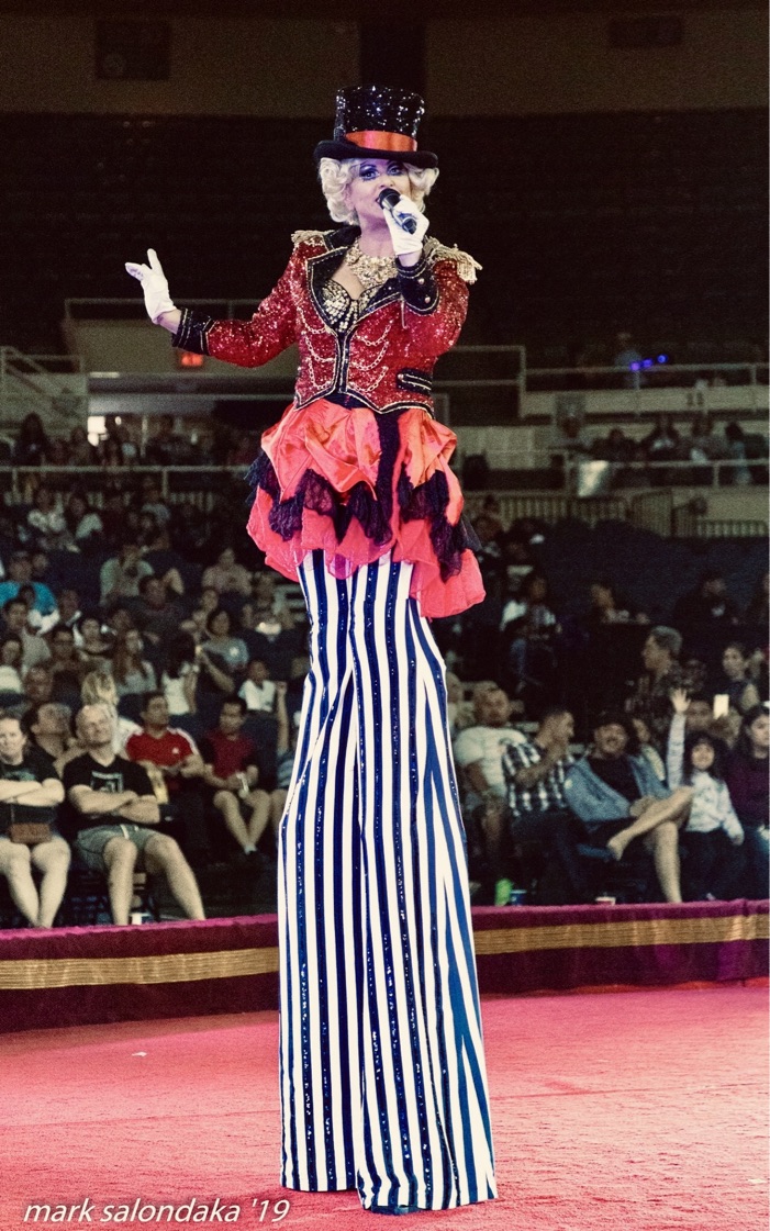 Miss Molly speaking into a microphone on stilts in a circus ring