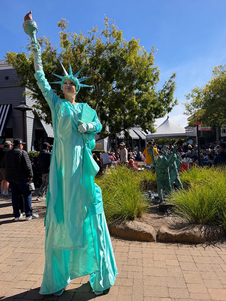 Miss Molly on stilts in a Statue of Liberty costume, holding a torch aloft